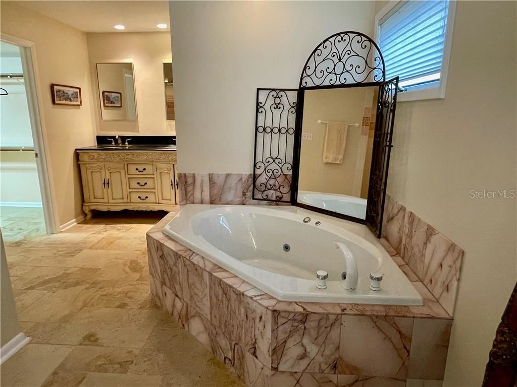 warm and cozy master bathroom with tub surrounded by marble and high window above tub, sink and mirror in the background