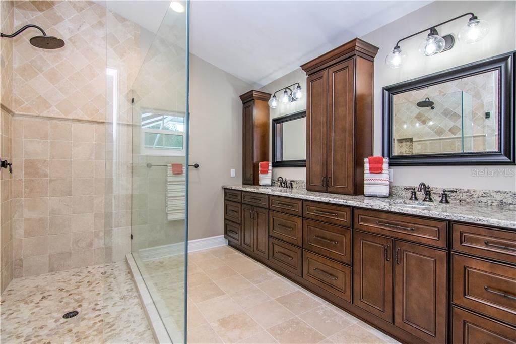 stunning brown bathroom with large glass shower with tile. big dark brown cabinets and drawers with two large mirrors and lots of space