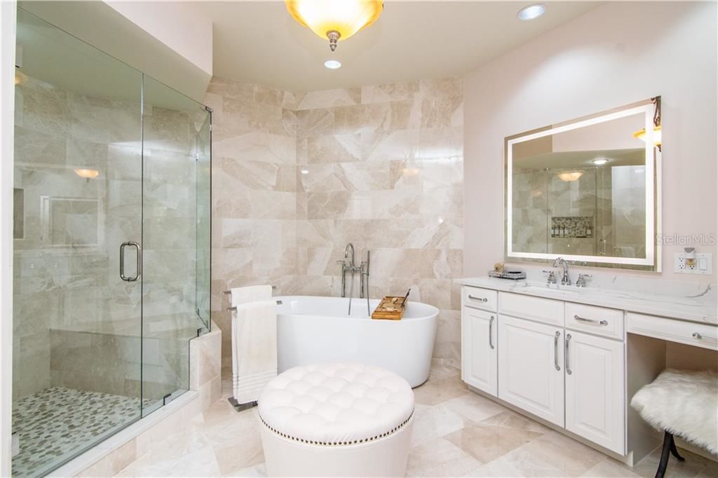 stunning bathroom with tile floors and wall behind bathtub. large glass shower. white cabinet drawers and light-up mirror.