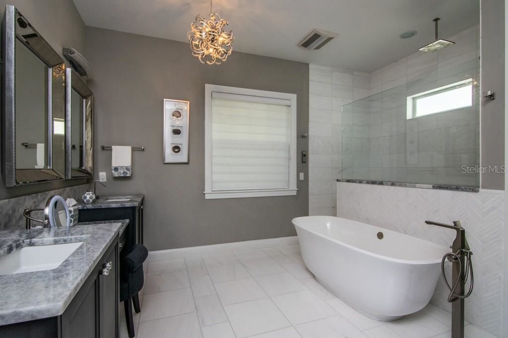 gray master bathroom with marble counter top 3 mirrors abd a large tub in front of a glass shower