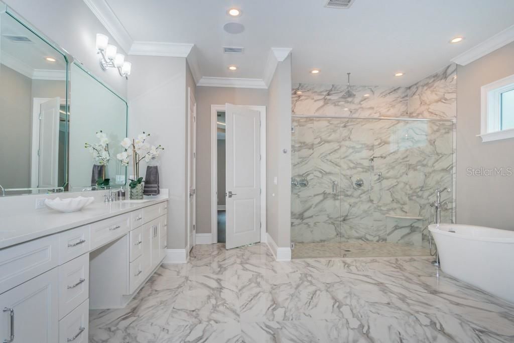 lovely master bathroom with a large mirror and white drawers and vanity, marble floors and shower with glass doors and a large white soaking tub