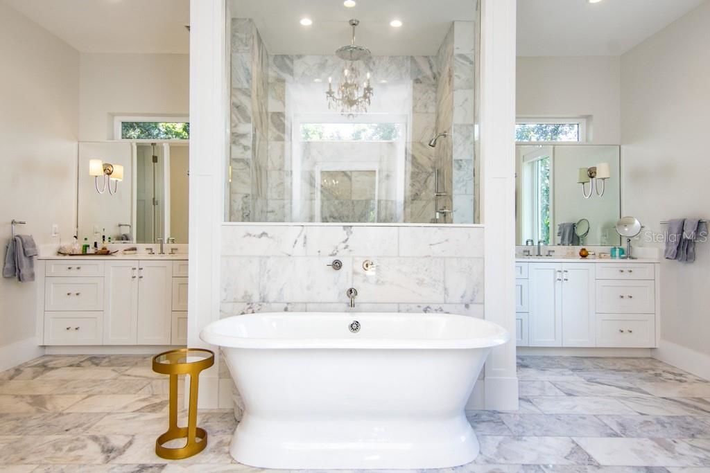 beautiful white dazzling master bathroom with a soaking tub in the center of the room with a chandelier above, behind is a glass shower and sink to either side 